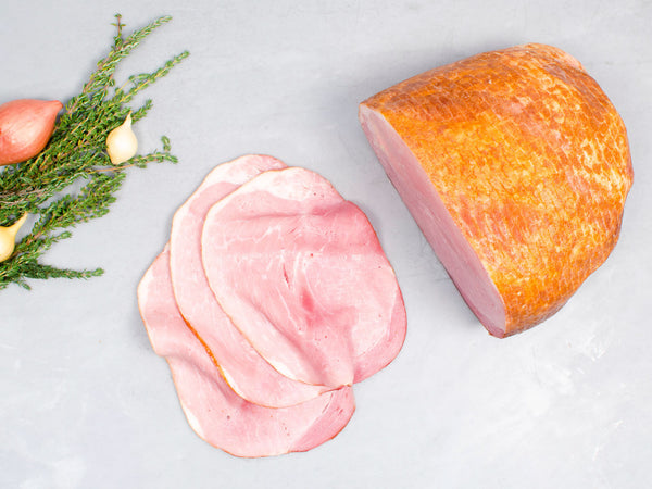 Sliced heritage ham with a whole piece and herbs on a light gray background