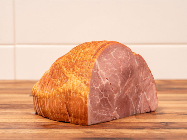 A sliced cured ham or prosciutto crudo showing both the golden-brown smoked skin and the pink marbled meat interior, resting on a wooden surface against a light tiled background.