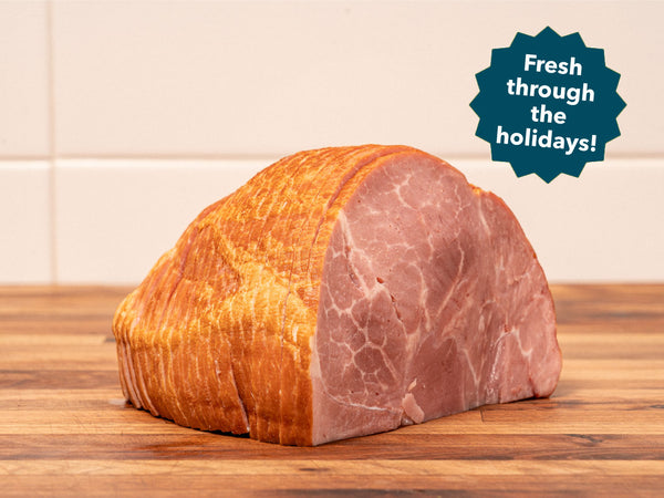 A sliced ham or roast with a golden-brown bark crust sits on a wooden surface against a light tiled wall. A blue burst badge in the top right reads 'Fresh through the holidays!'.