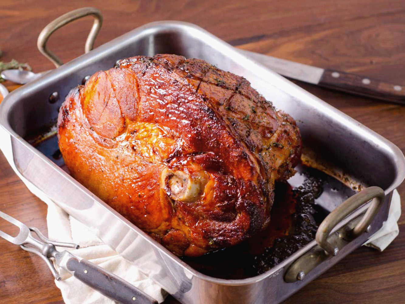 A roasted ham in a stainless steel roasting pan on a wooden table, with carving tools nearby.
