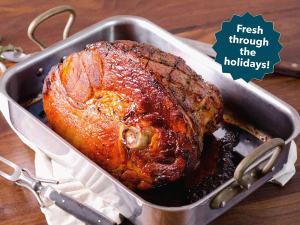 A roasted ham in a stainless steel roasting pan on a wooden table, with a teal badge that reads 'Fresh through the holidays!' in the upper right.