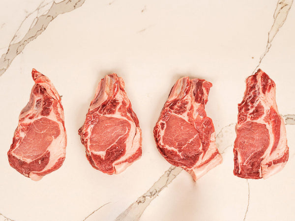 Four raw bone-in lamb chops arranged in a row on a light beige marble surface.