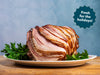 A sliced spiral ham on a white platter with green parsley, set on a wooden table against a textured blue background. A dark blue starburst badge in the top right reads 'Fresh for the holidays!'.