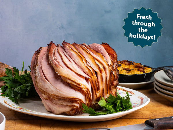 A sliced spiral ham on a white plate with parsley garnish, set on a wooden table with a blue wall background and a teal badge that reads 'Fresh through the holidays!'.