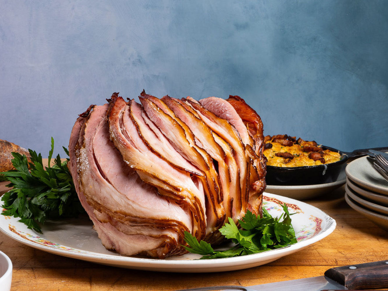 A sliced spiral ham with a golden-brown glazed exterior, served on a white plate with parsley garnish, set on a wooden table with a blue textured background.