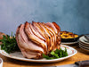A sliced spiral ham with a golden-brown glazed exterior, served on a white plate with parsley garnish, set on a wooden table with a blue textured background.